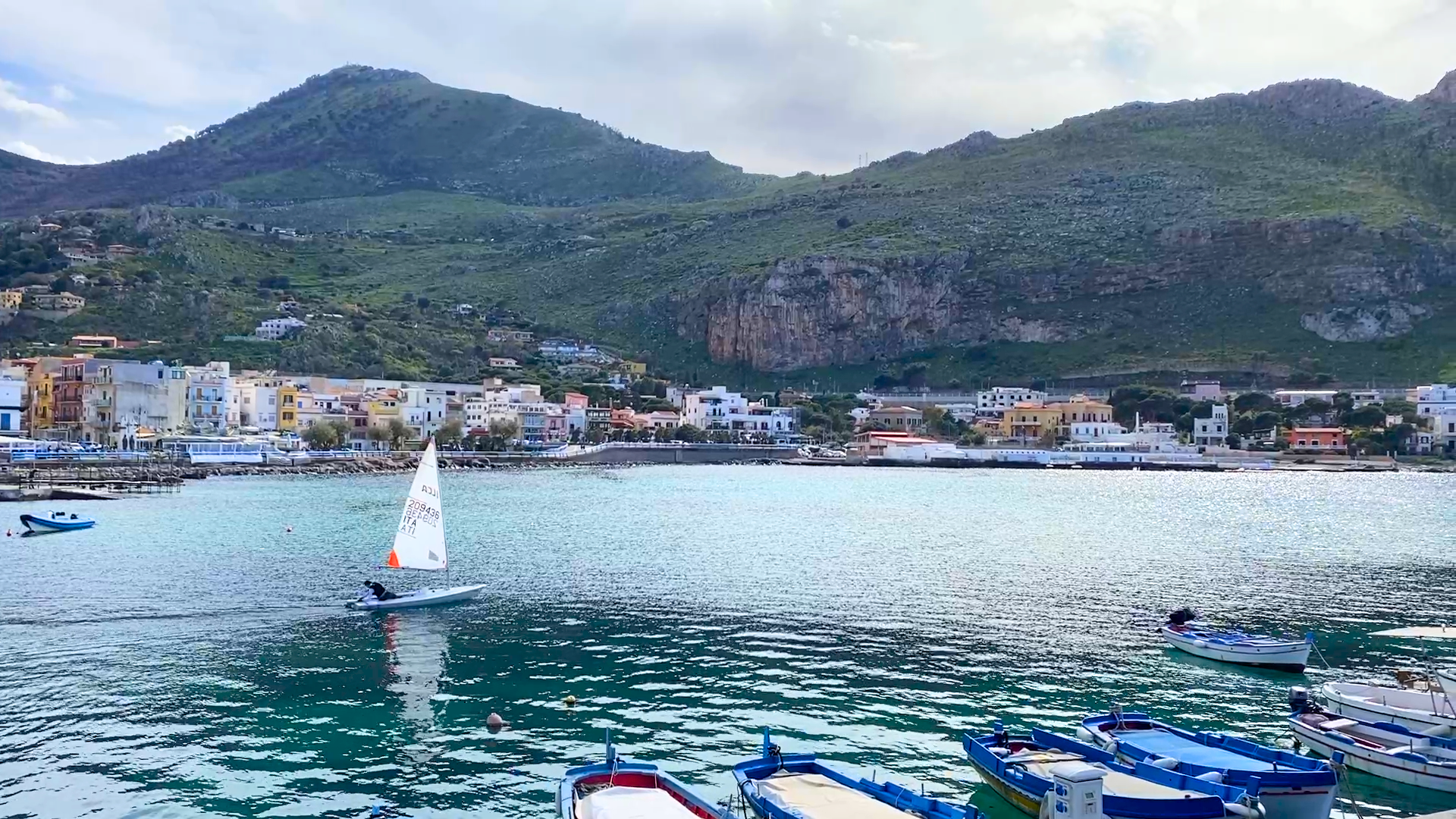Small sailboat gliding through the harbor of Sferracavallo, Sicily, with colorful boats and mountains in the background during spring in Sicily