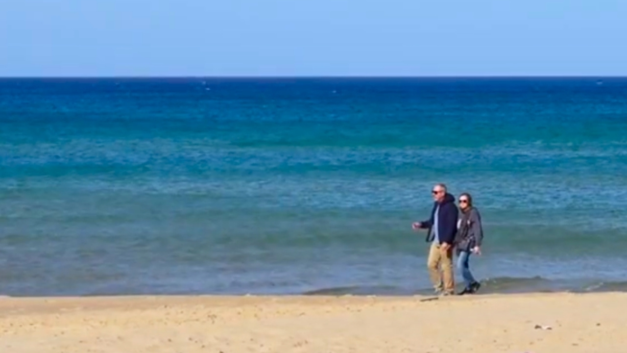 Joe and Diana walking along a quiet beach living in Sicily with calm blue water